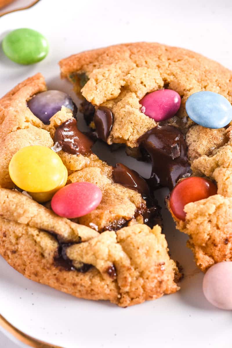 Cookie shown broken in half, you can see the melted dark chocolate chips and the colourful smarties on top of the cookies. 