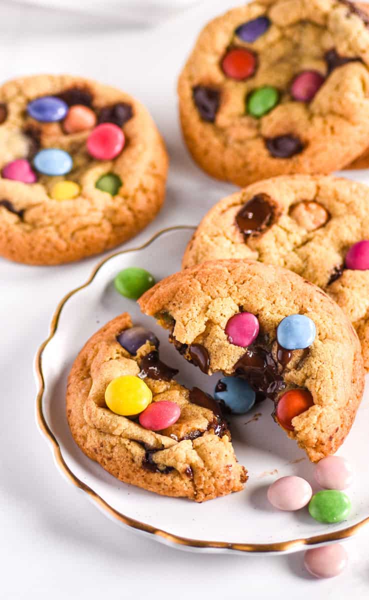 Smarties Cookies shown on a plate and more in the background. There are smarties scattered around the cookies. 