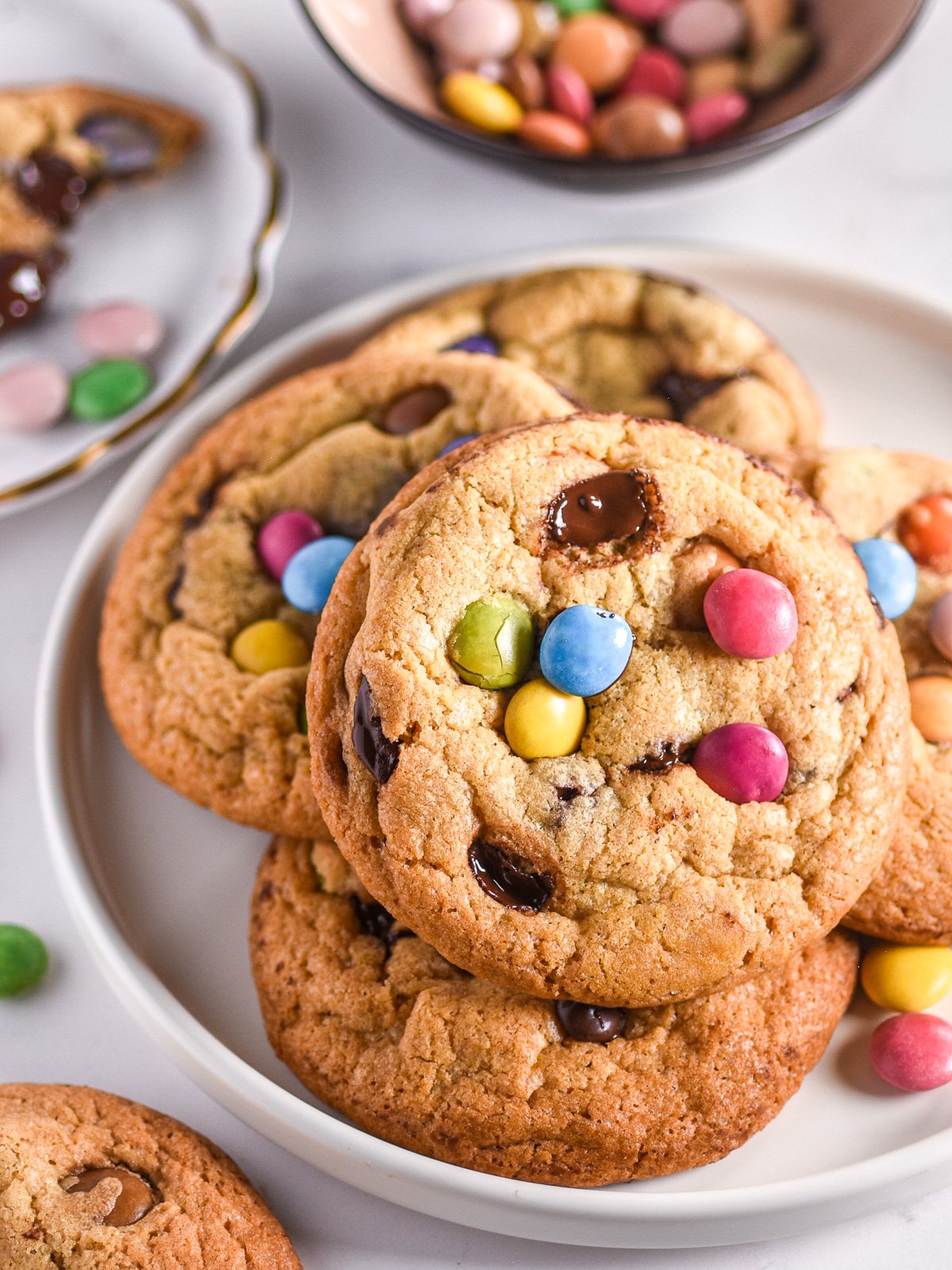 Smarties Cookies shown stacked on a plate, there is a bowl of smarties in the background. 