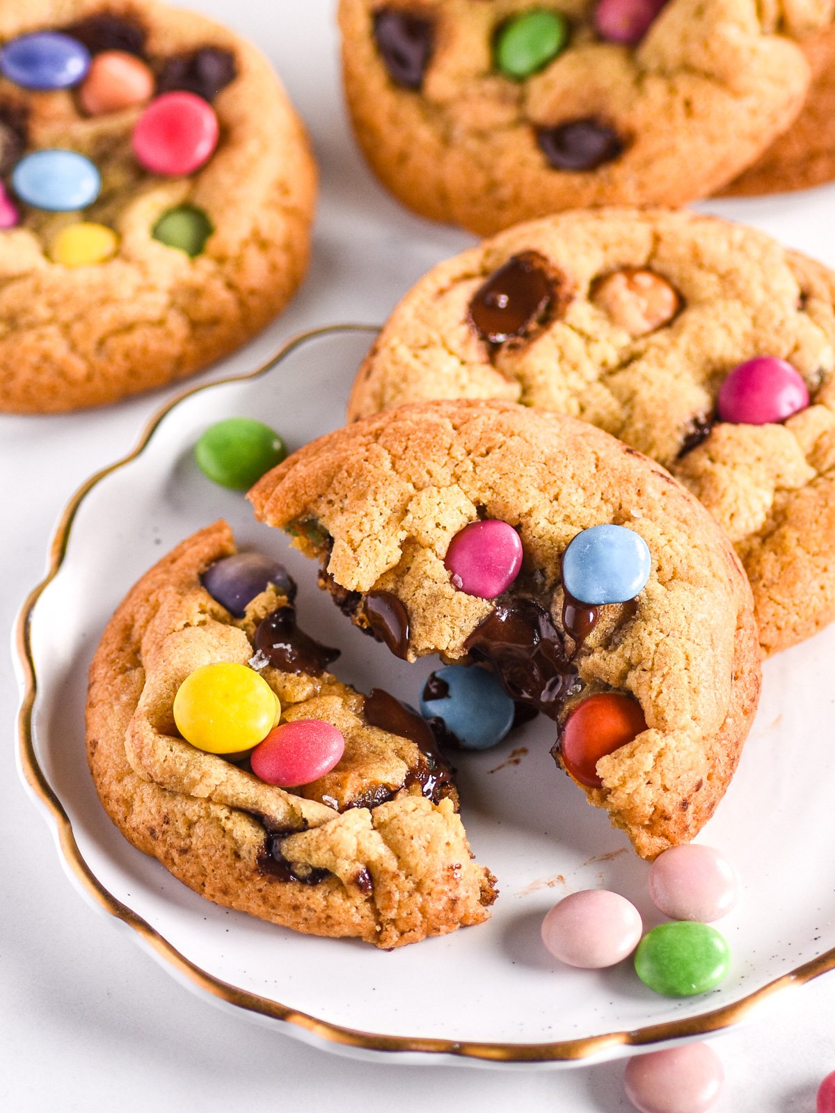 Smarties Cookies shown on a white plate with smarties scattered around. The cookie in the fron tis broekn in half. 