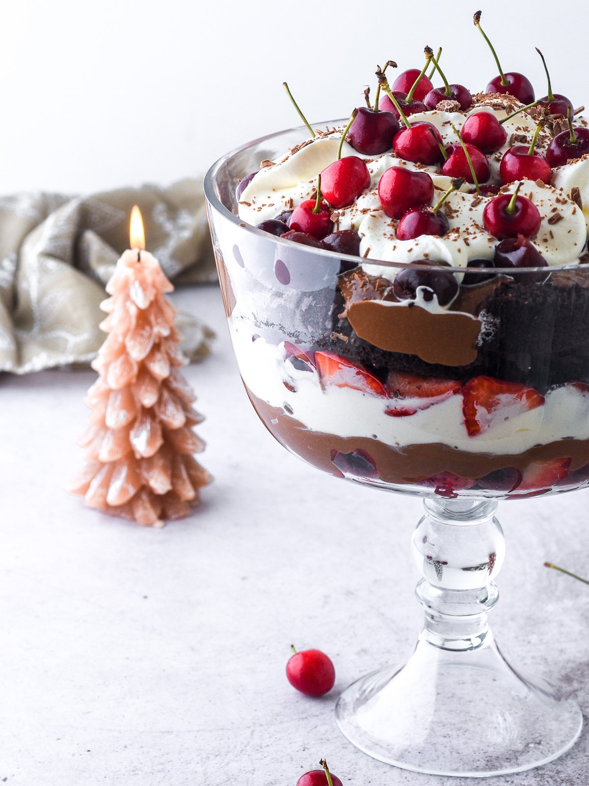 Black Forest Trifle shown in a trifle dish with a stand, there is a Christmas tree candle in the background.