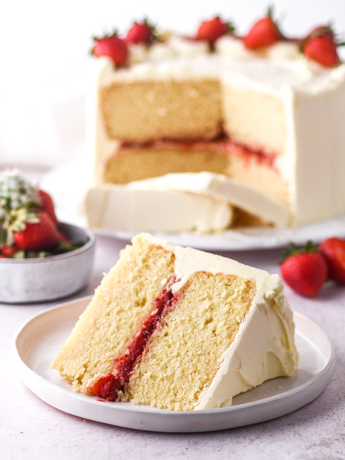 Image showing a slice of strawberry and vanilla cake, there is the whole cake in the background.