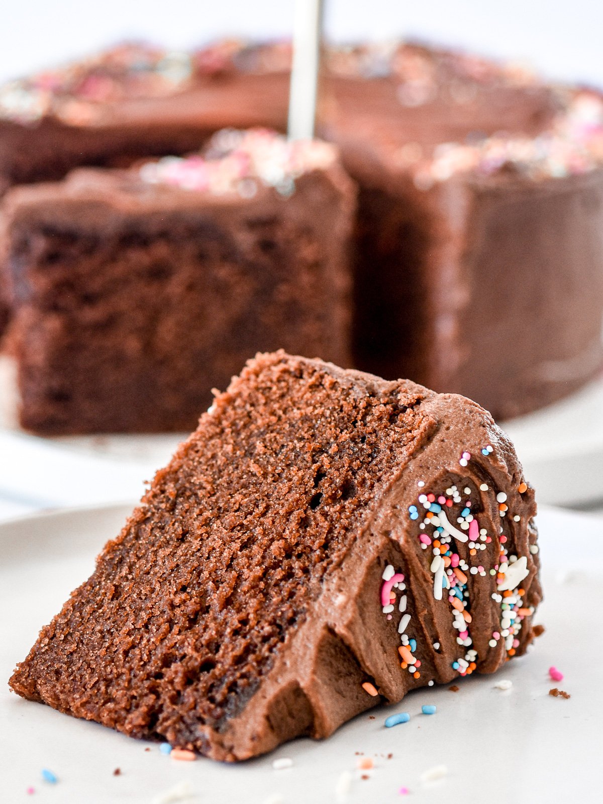 Image showing a slice of chocolate cake with sprinkles, and the whole cake in the background.