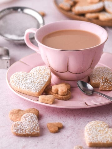 Pink tea cup on a saucer with heart shaped biscuits on the saucer and counter top.