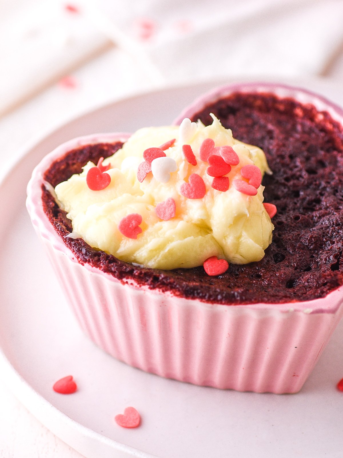 Image shows a heart shaped ramekin filled with a baked red velvet mug cake topped with a dollop of cream cheese frosting.