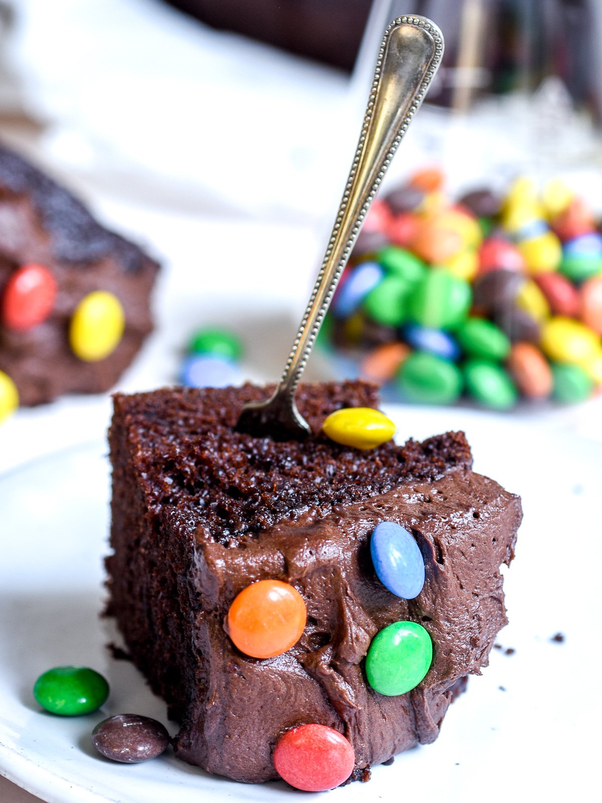 Image showing a square slice of chocolate cake with chocolate cream cheese frosting decorated with smarties on a plate with a fork through the slice.