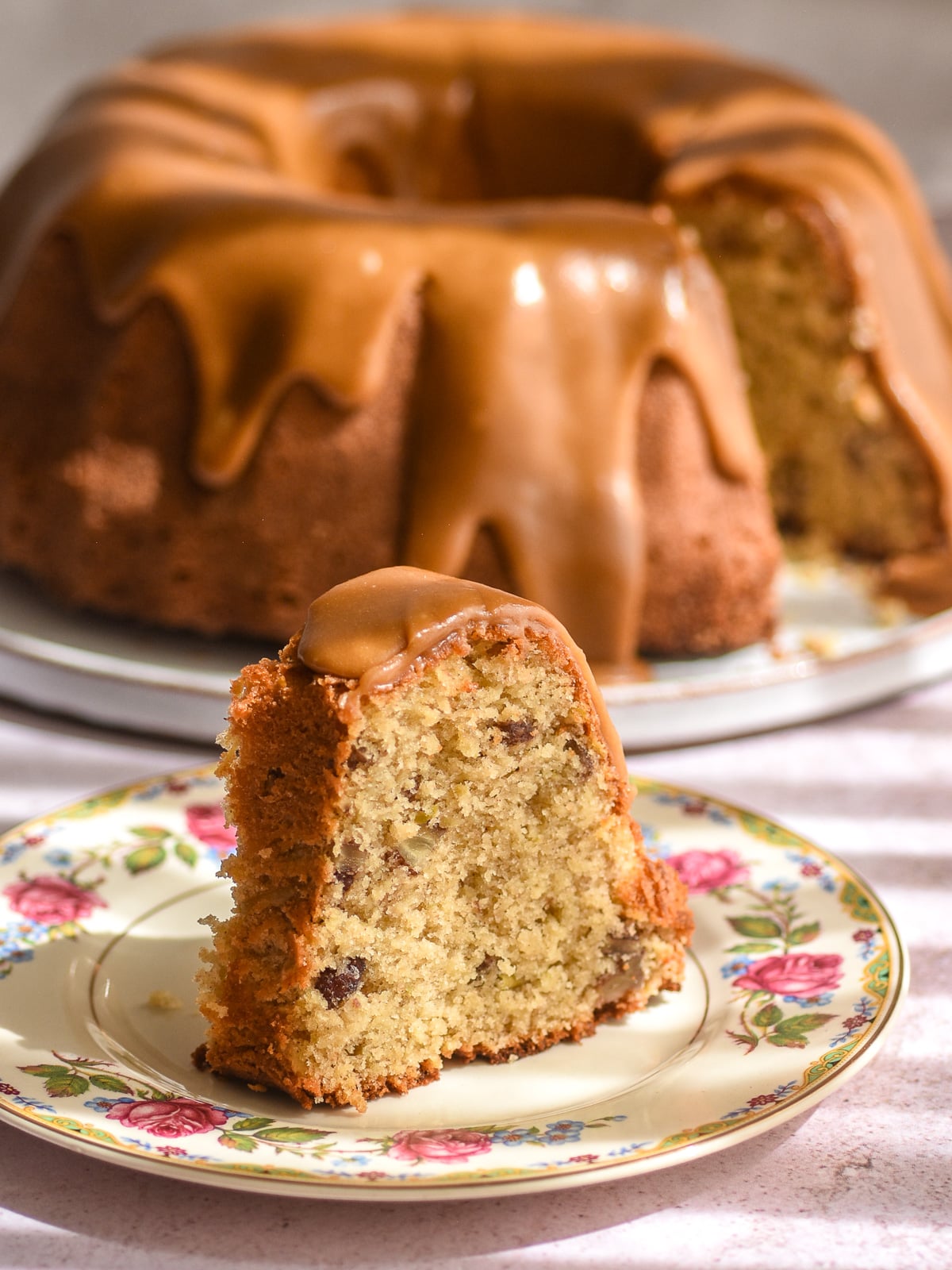A slice of apple pecan cake with caramel pecan sauce on a plate, with the rest of the bundt cake in the background.